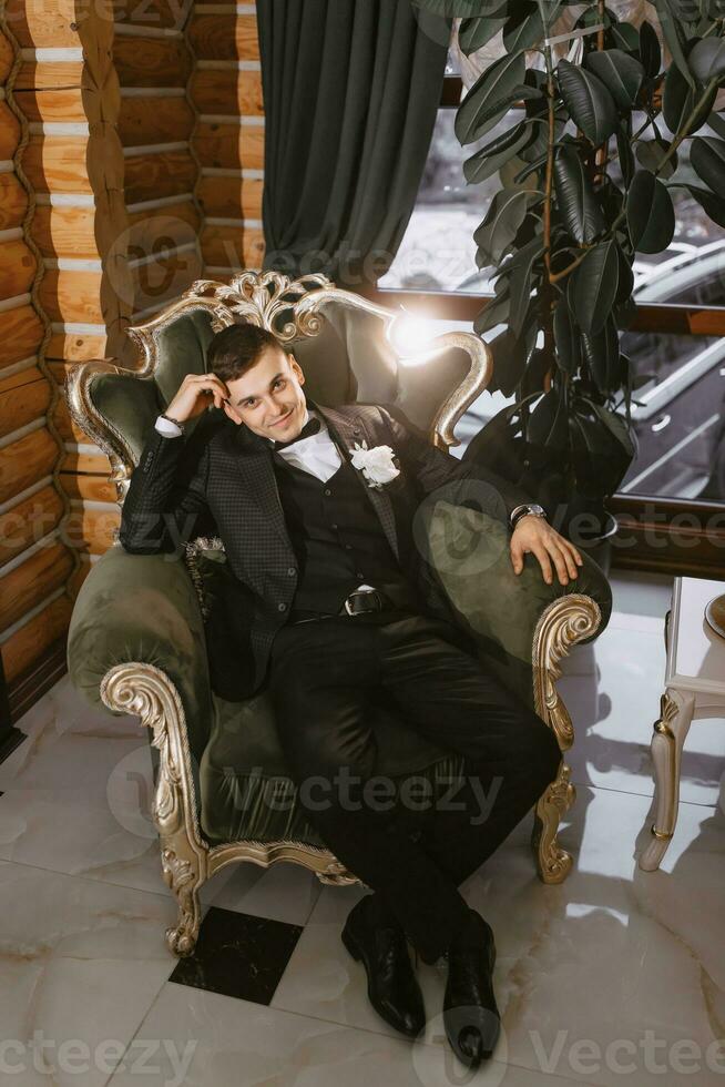 portrait of positive young groom in black suit with white shirt smiling and sitting on comfortable dark green sofa in modern hotel room on wedding day photo