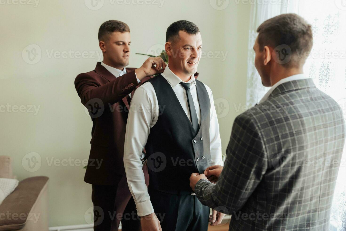 Photo of three men in classic suits. A handsome young man mends the shirt of another man, standing behind him. Business style. Stylish men