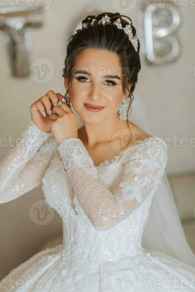 Beautiful young bride getting dressed before wedding ceremony at home. A brunette bride in a wedding dress wears earrings photo