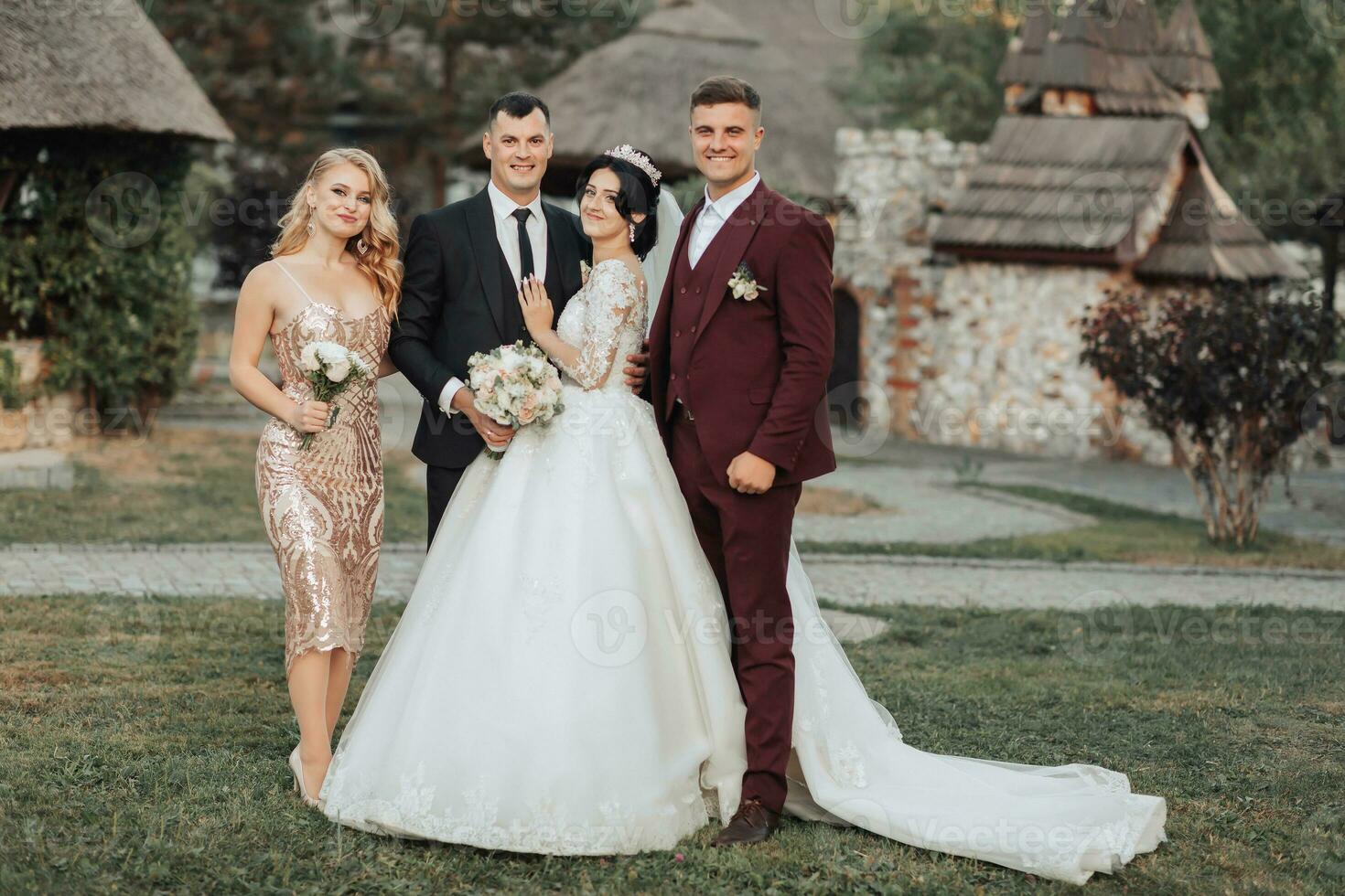 Front view of newlyweds walking hand in hand among friends. Girls in golden dresses hold bouquets, the bride in a voluminous dress, the groom and his friends in suits. Wedding in nature photo