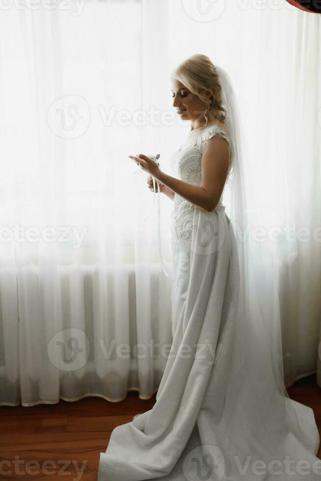 hermosa rubia novia retrato, participación Cordon de zapato en mano Boda maquillaje peinado, maravilloso joven mujer en blanco vestir a hogar. serie foto