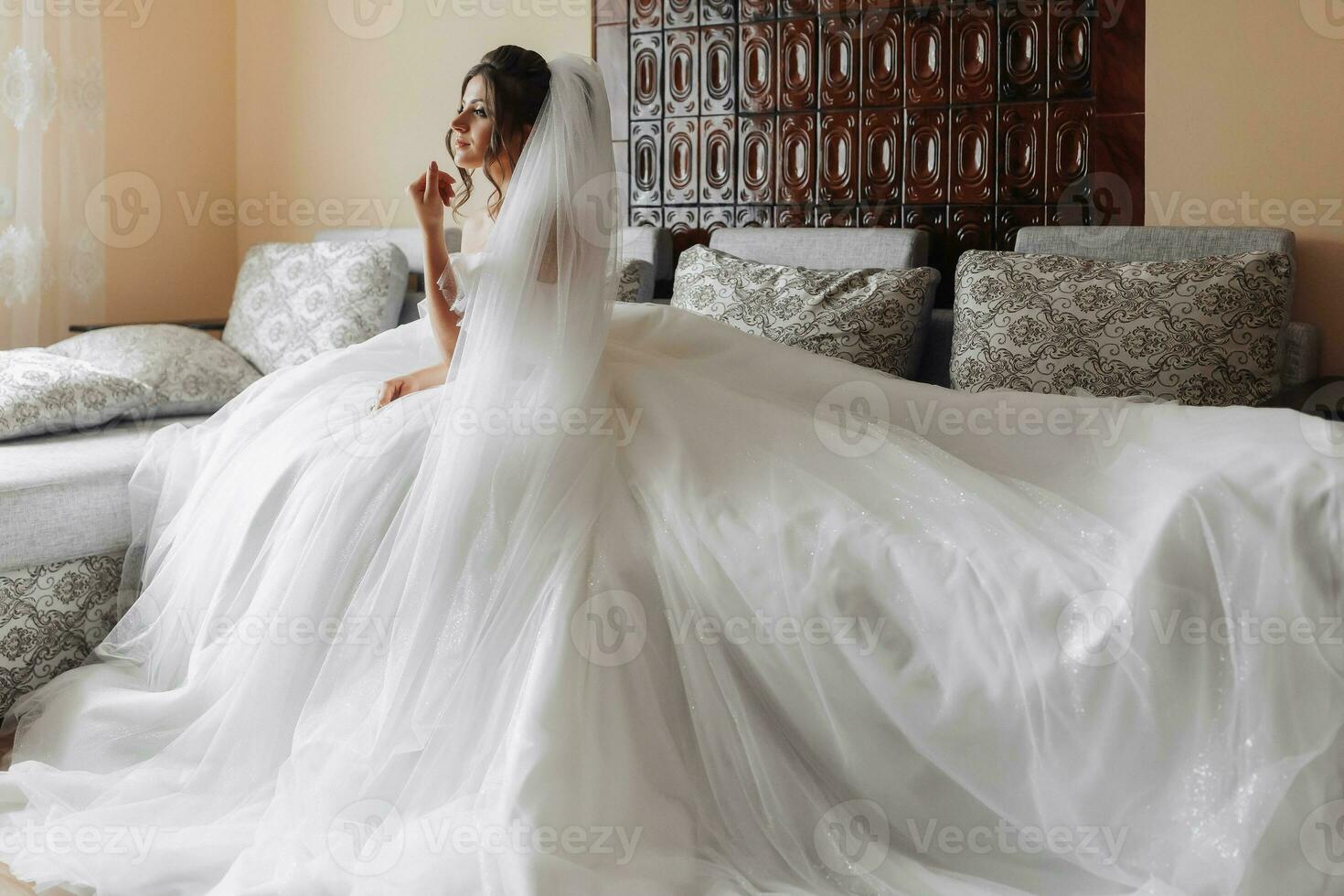 A brunette bride in a white dress poses while sitting on a gray sofa and holding her curls. Gorgeous makeup. Open shoulders. Beautiful hands. Long veil. Morning of the bride. wide angle photo