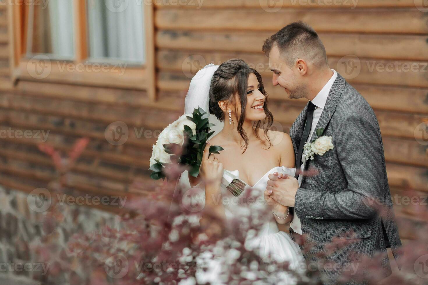 Portrait of the bride and groom in nature. A brunette bride in a long white off-the-shoulder dress and a veil and a stylish groom pose hugging near a mahogany tree. Beautiful curly hair and makeup. photo
