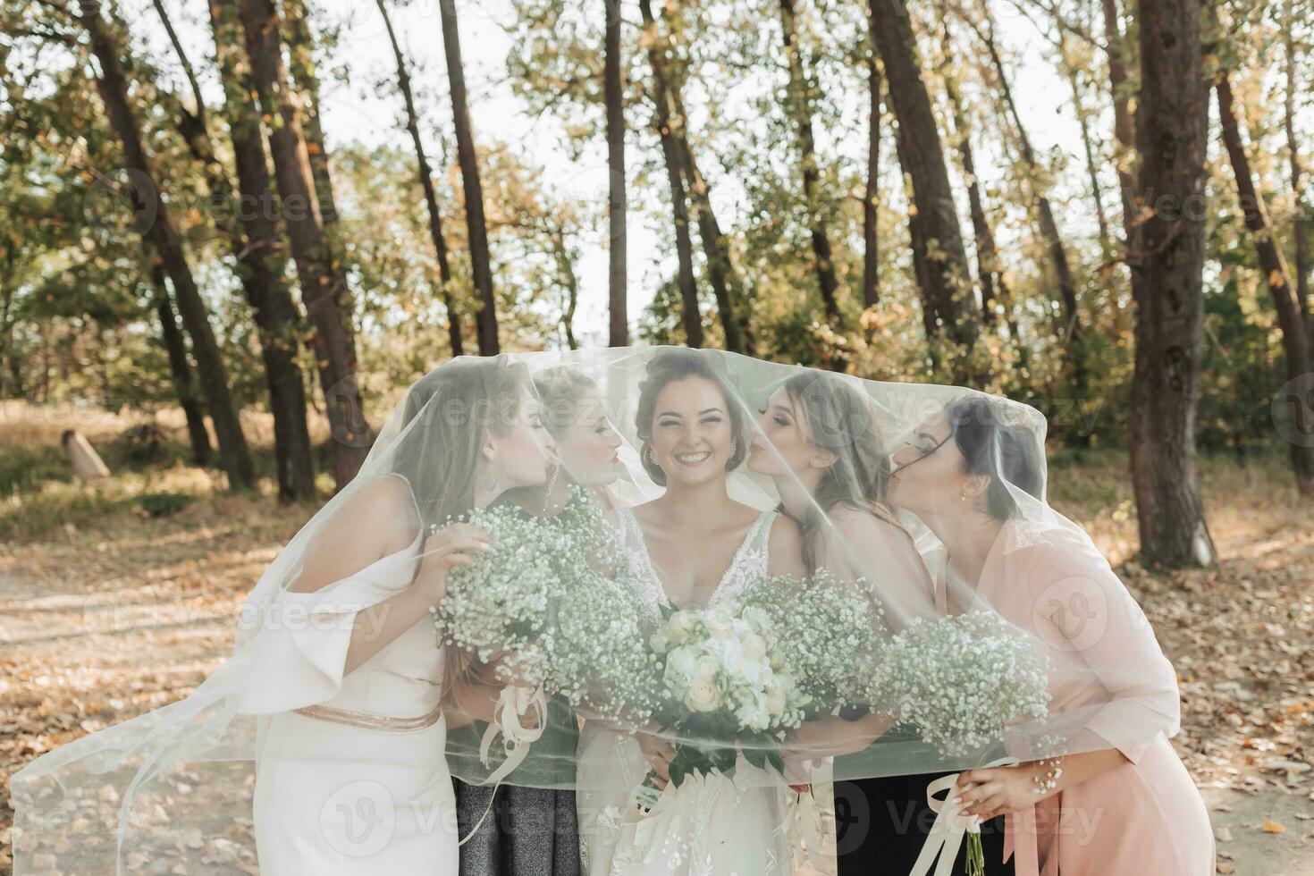 Wedding photo in nature. The bride and her friends are standing in the forest holding their bouquets, smiling, covered by the bride's veil. Wedding portrait. Emotions. Beautiful girls.