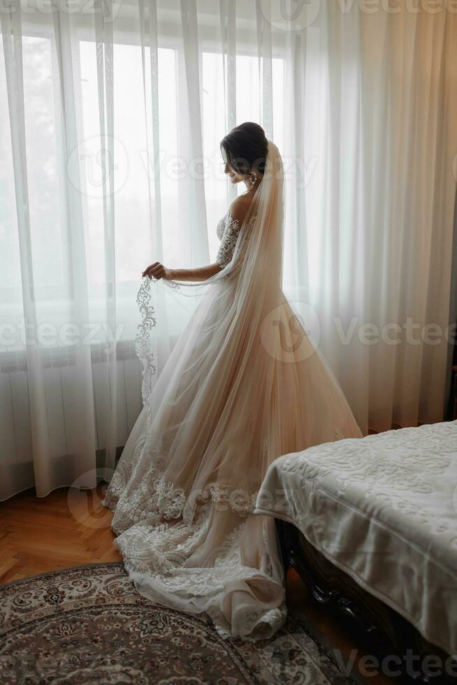 A brunette bride in a white dress and a long veil poses standing by the window. Beautiful hands. French manicure. Beautiful hair and makeup. Daylight photo