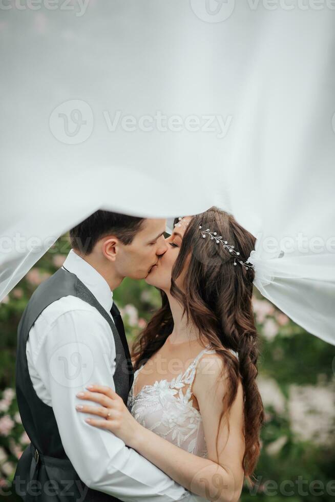 Happy young couple. Wedding portrait. The bride and groom tenderly kiss under a veil against the background of a blooming bush. Wedding bouquet. Spring wedding photo