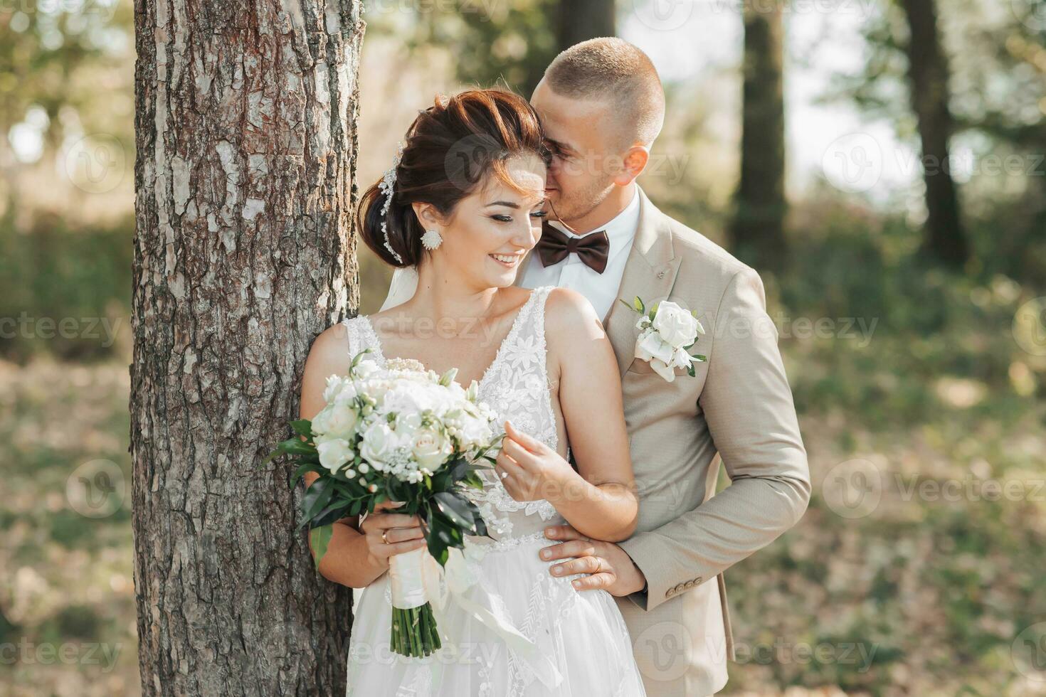 Boda foto en naturaleza. el novia y novio son en pie cerca un árbol, el novio abrazos su amado desde detrás y Besos su, ella sonrisas atentamente. retrato. verano Boda