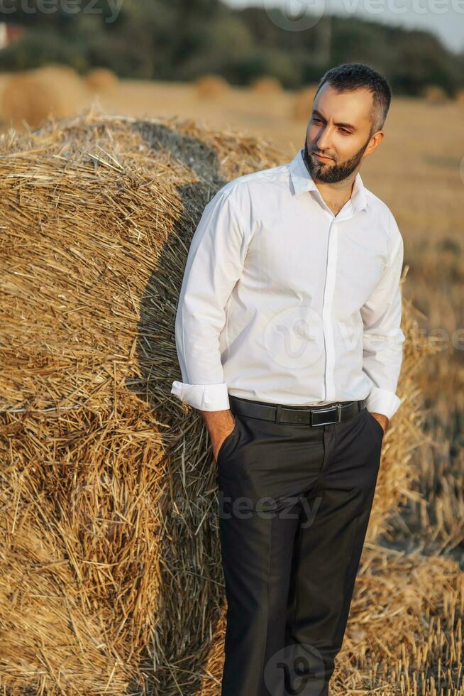 Boda retrato foto. un elegante novio en un blanco camisa poses cerca un bala de heno con su manos en su bolsillos barbado hombre. estilo. foto