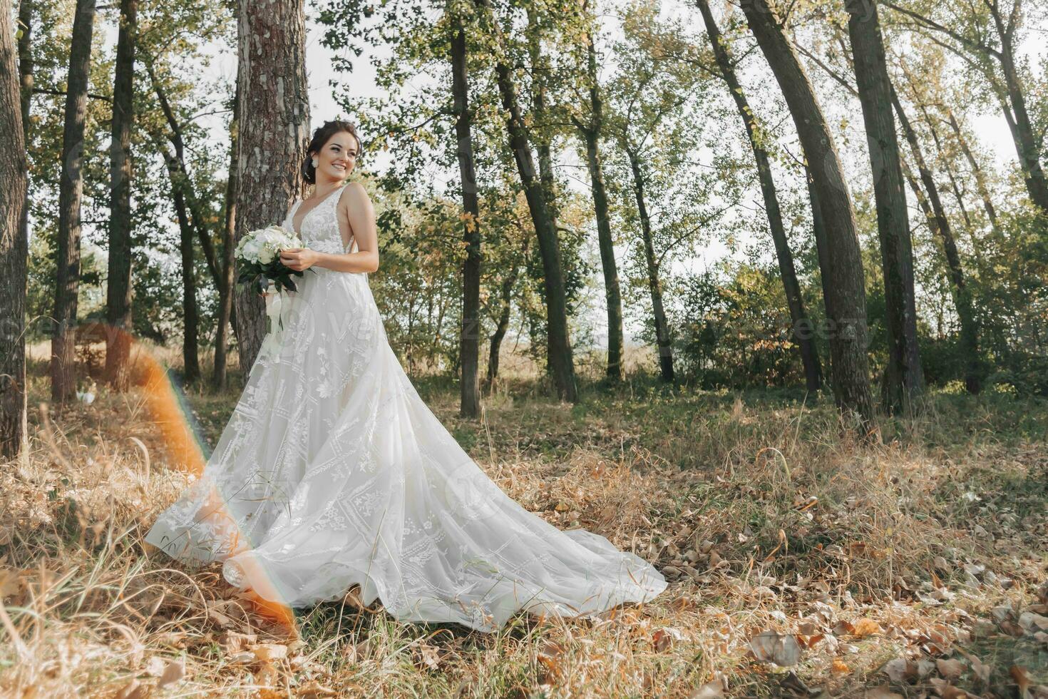 Wedding photo in nature. The bride is standing in the forest. The bride in a beautiful dress with a long train, holding her bouquet of white roses, smiling sincerely at the camera. Portrait