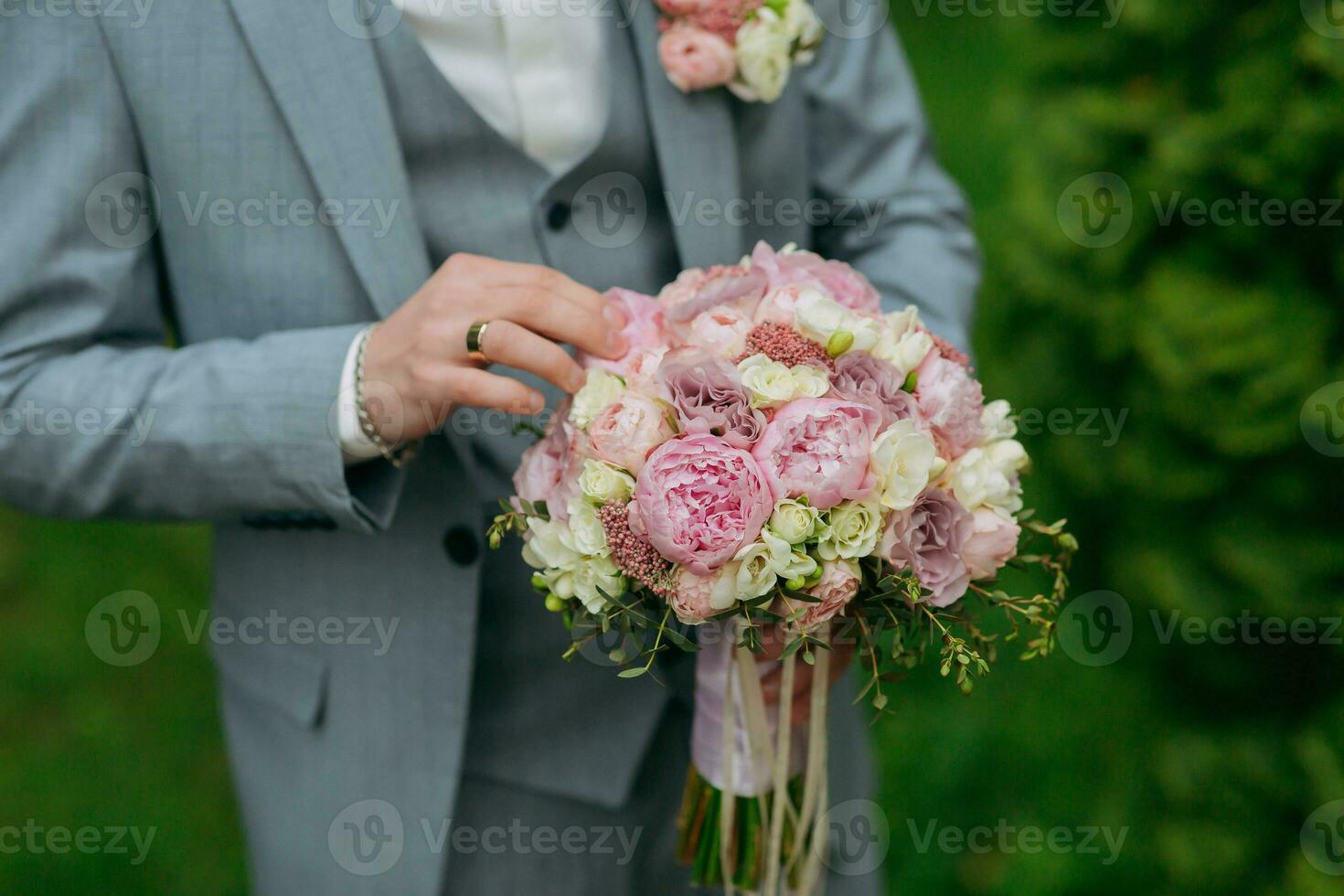 Portrait of the groom standing against the background of green trees and holding a wedding bouquet of pink peonies. Side view. Stylish groom. Fashion and style. photo