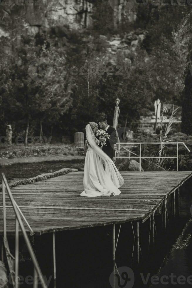 Groom in a black suit and white shirt dancing on a pier with a bride in a white wedding dress, near a lake and rocks. Black and white photo