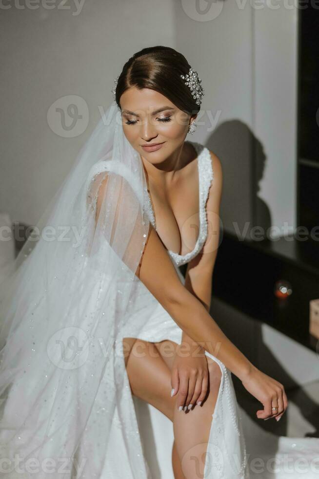 Young bride in a white dress sitting on a sofa with a long veil, portrait photo. Preparation for the wedding ceremony. Accessories for newlyweds photo