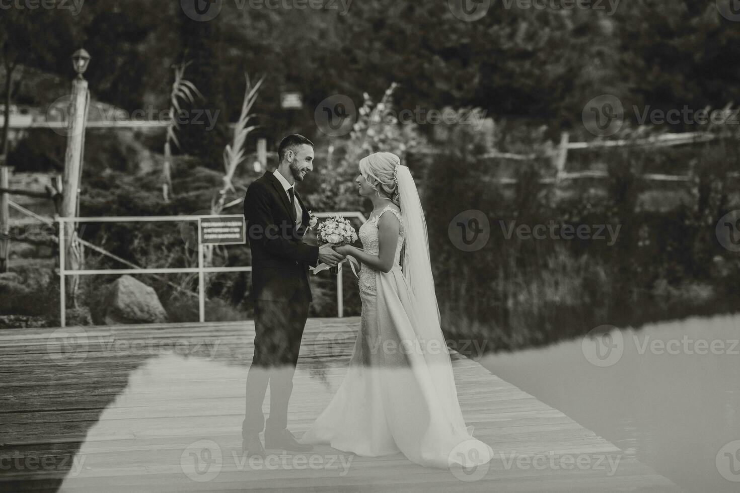 The groom in a black suit and white shirt gently touches and holds the hand of the bride in a white wedding dress, standing near a lake and stones photo