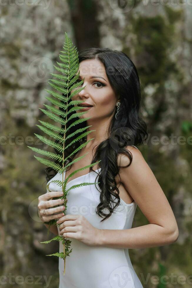 A girl with long black curly hair, standing in a white silk dress in a spring forest against a large tree, holding a fern branch in her hands photo