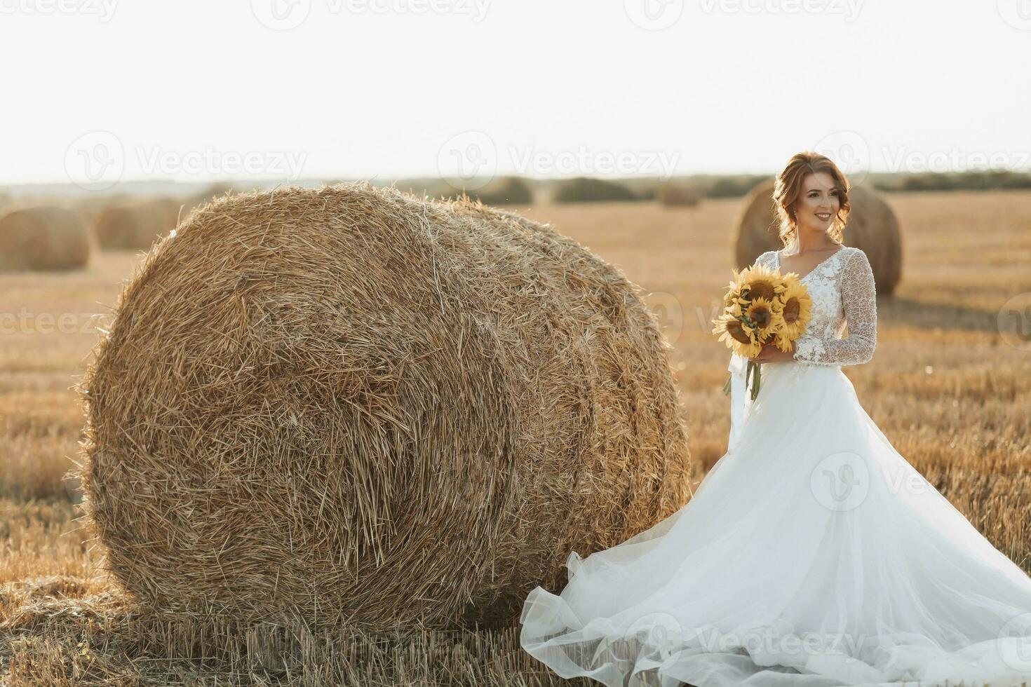 Wedding portrait. A red-haired bride in a white dress is standing near a bale of hay, holding a bouquet of sunflowers, against the background of a field. Beautiful curls. Sincere smile. Elegant dress. photo