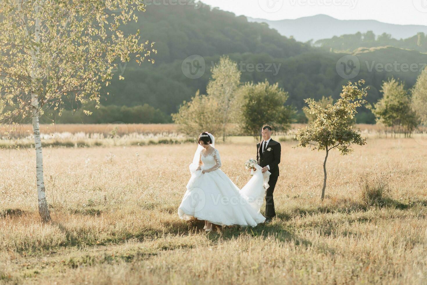 a bride and groom walking against a background of trees and a field. The bride is in front of the groom, the groom is holding the train of the dress behind her. Wedding photo in nature