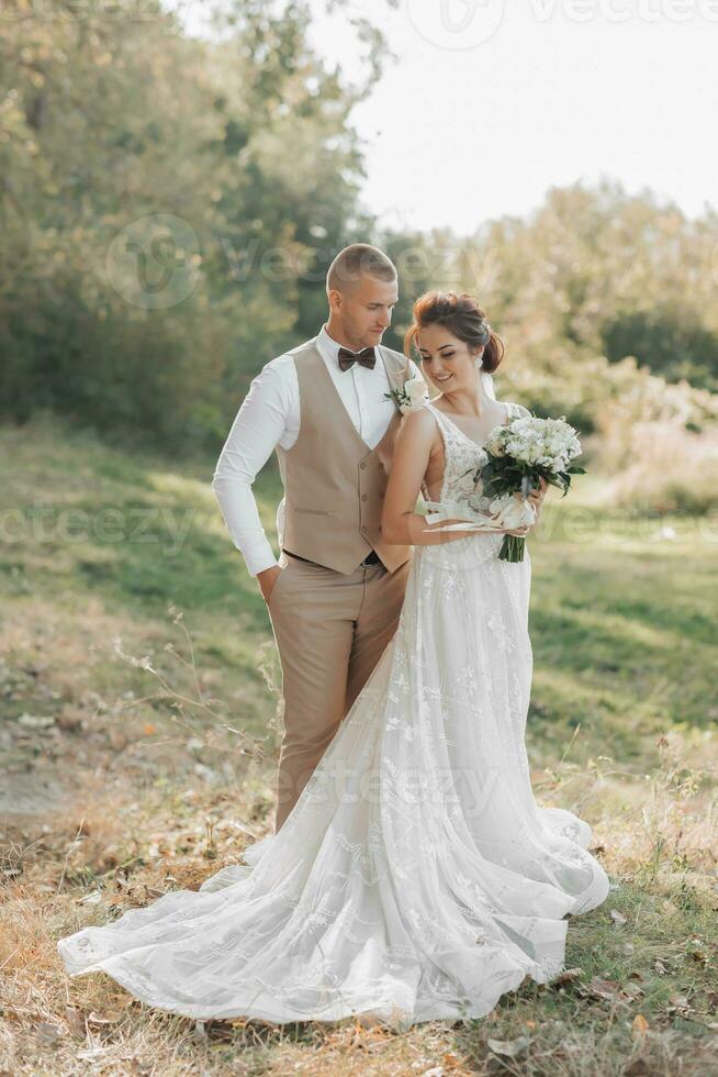 Wedding photo in nature. The groom hugs his beloved from behind and looks at her, the bride holds a bouquet and looks to the side. Long train of the dress. Portrait. Summer wedding.