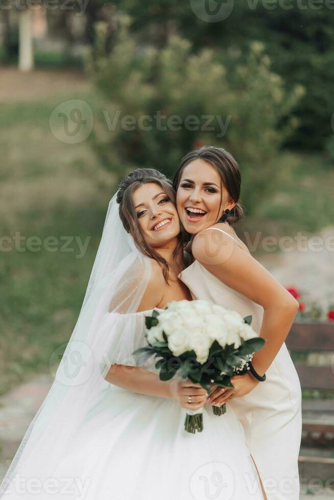 Wedding photo in nature. A brunette bride in a white long dress and her friend in a nude dress are hugging and smiling and posing. Young women. emotions