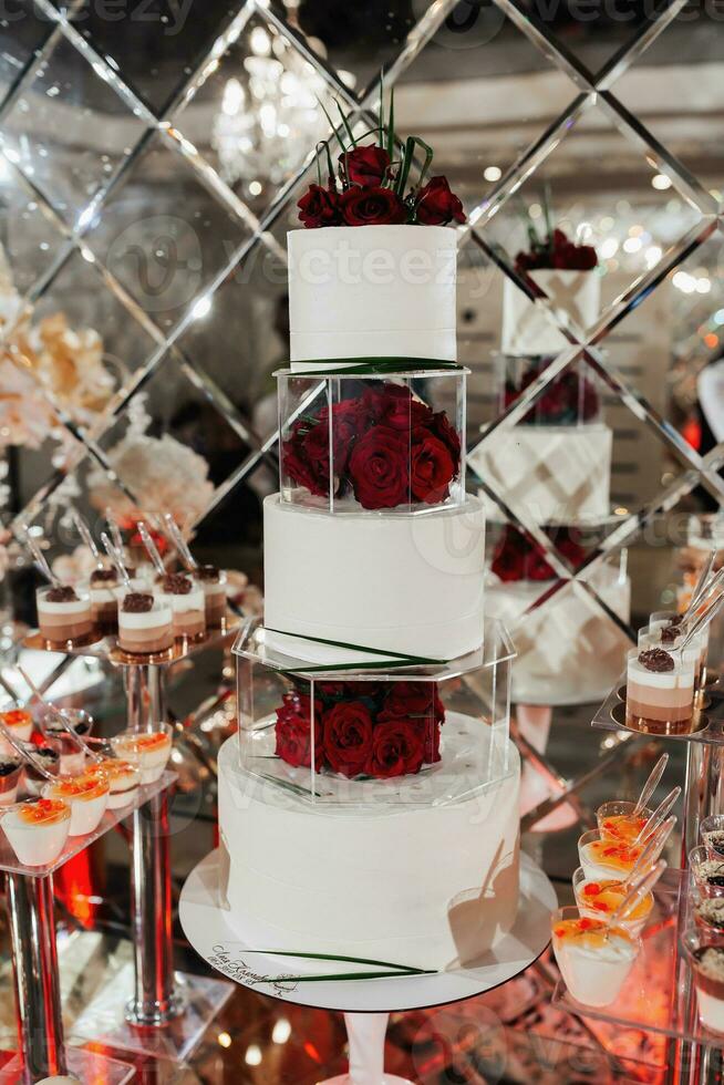 The white wedding cake is decorated with red roses. A delicious wedding. Candy bar for a banquet. Celebration concept. Fashionable desserts. A table with various sweets, candies. photo
