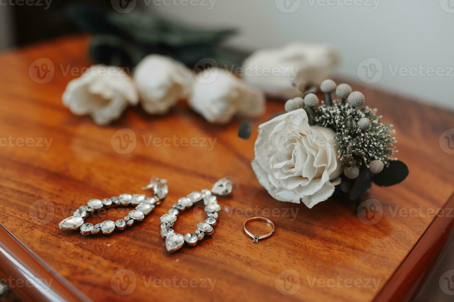 Photo of details at the wedding. Wedding ring with diamond and bride's earrings, boutonniere stands on wooden background. Beautiful light. Fashion and style