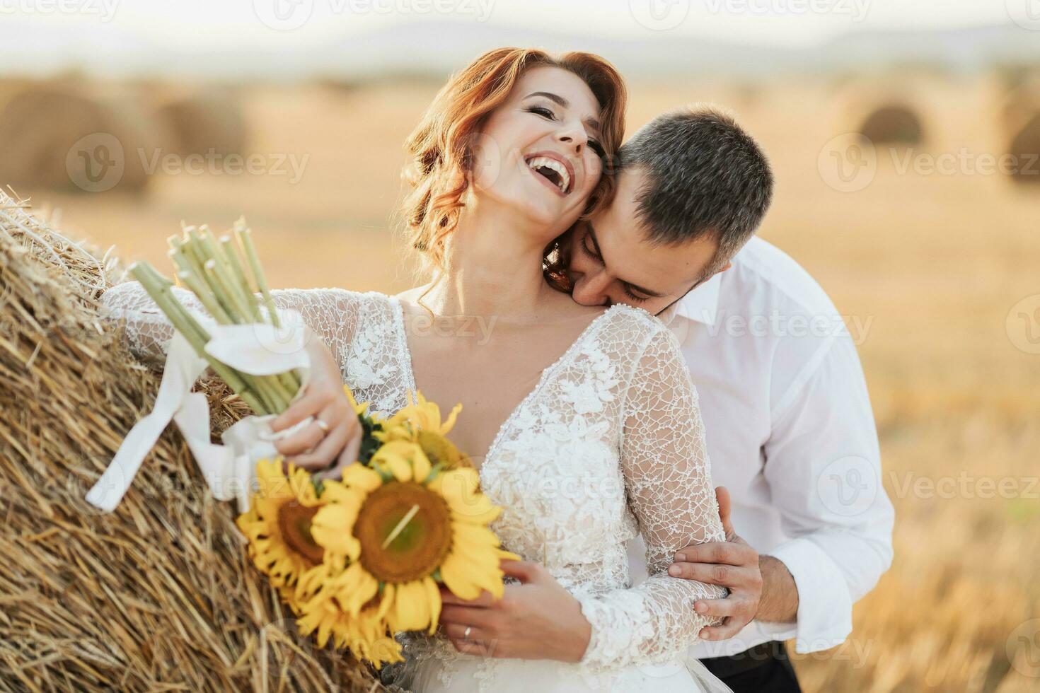 Wedding portrait of the bride and groom. The groom, tearing his shirt, stands behind the bride, near a bale of hay. Red-haired bride in a long dress with a bouquet of sunflowers. Stylish groom. Summer photo