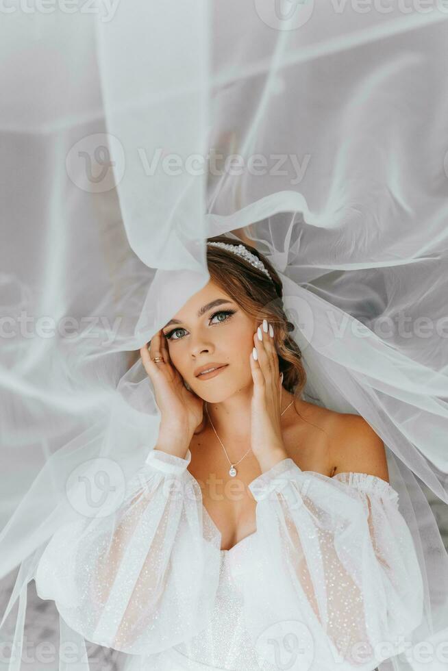 Beautiful curly brown-haired bride in a white dress poses for a photographer, standing under a veil in a beautiful dress with sleeves. Wedding photography, close-up portrait, chic hairstyle. photo