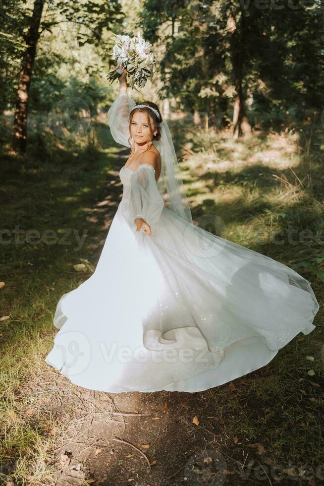 un hermosa novia en un blanco vestir con un largo velo es dando vueltas con un ramo de flores en el parque. foto desde el atrás. joven mujer, Arte foto, boda, novia, abierto espalda. ligero.