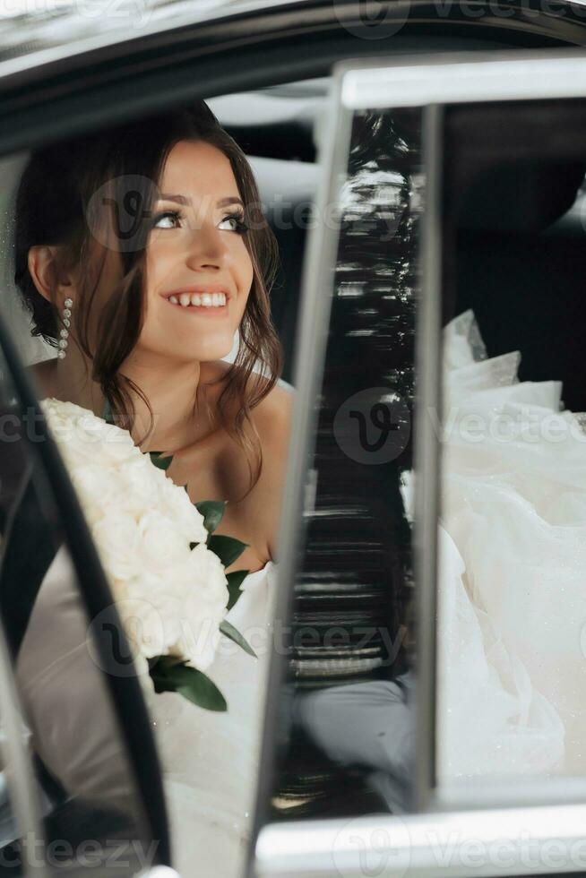 A brunette bride, smiling sincerely, sits in a black car on her wedding day with a bouquet of white roses. Portrait of the bride. Beautiful curls. Beautiful makeup and hair. A luxurious white dress photo