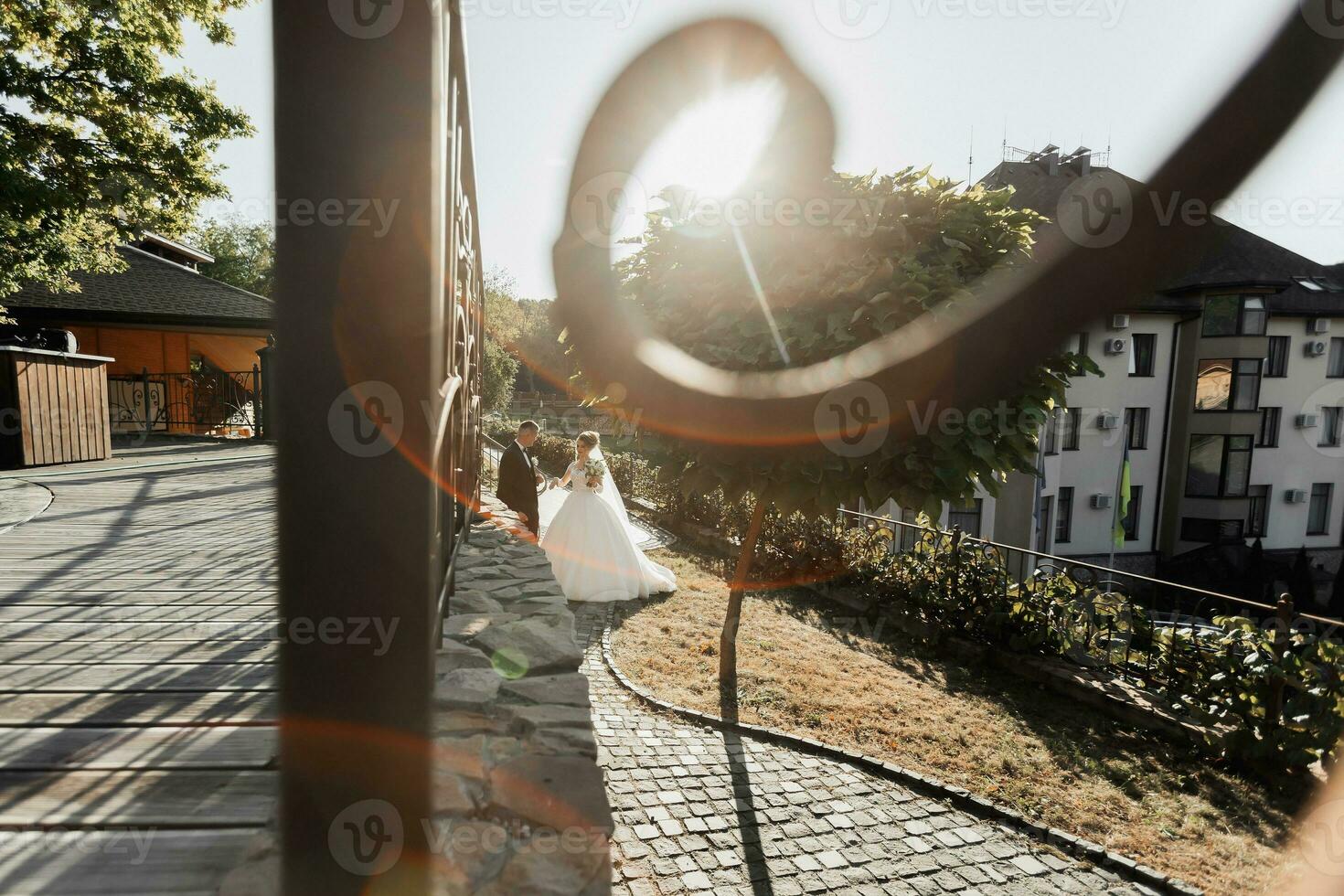 Wedding portrait. The groom in a black suit and the blonde bride are walking holding hands near a stone wall under a tree. A white, long dress in the air. Sun rays in the photo