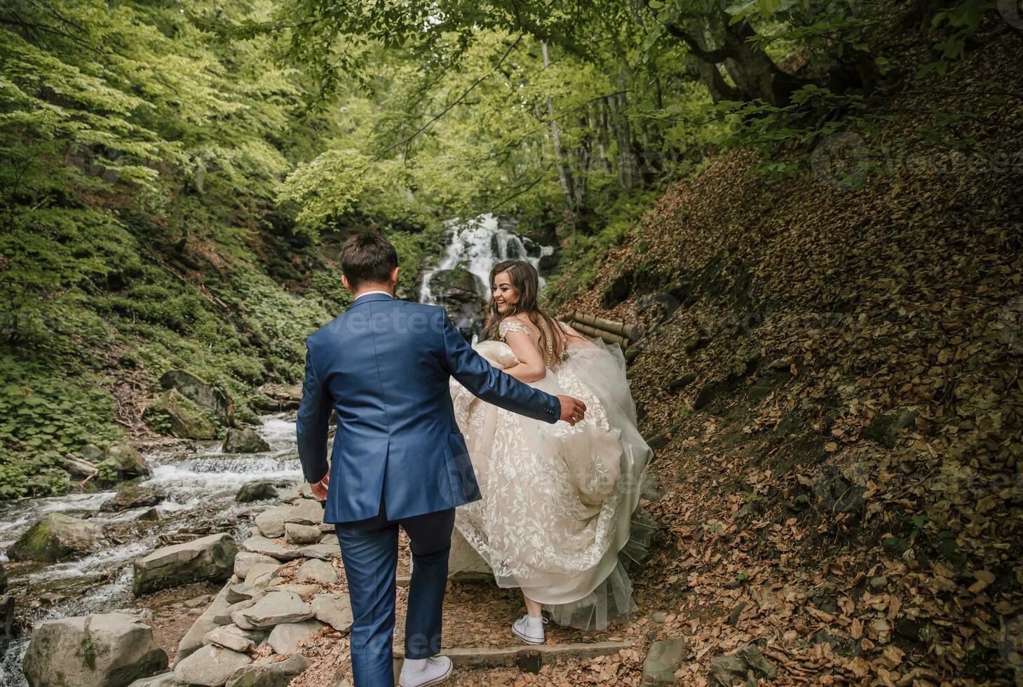Happy wedding couple running to the waterfall. The bride smiles sincerely. Wedding photo session in nature. Photo session in the forest of the bride and groom.