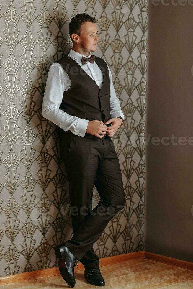 Confident elegant business man is buttoning up a brown vest while looking out the window. a full-length portrait in the room. photo