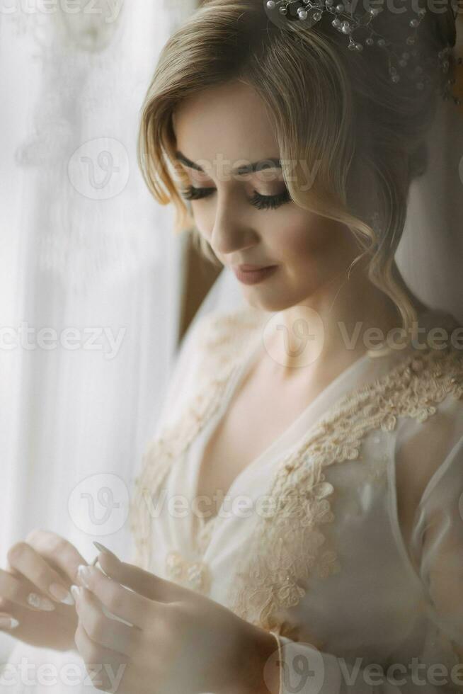 A bride with an elegant hairdo and tiara, in a robe, stands holding her ring and looking at it. Beautiful makeup. Wedding photo. Blonde photo