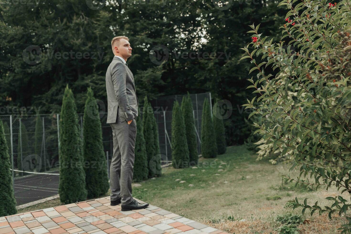 A man in a gray suit poses in nature with his hands in his pockets, his shoulders turned to the camera. Side view. A stylish watch. Men's style. Fashion. Business photo