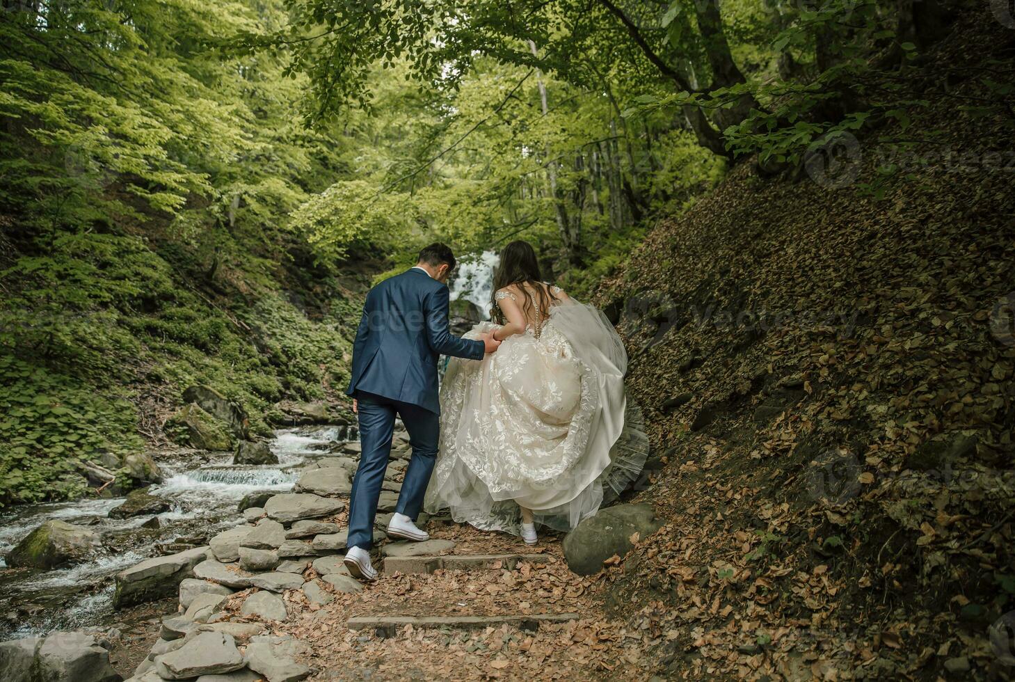 Happy wedding couple running to the waterfall. Bride and groom. Photo from the back. Wedding photo session in nature. Photo session in the forest of the bride and groom.
