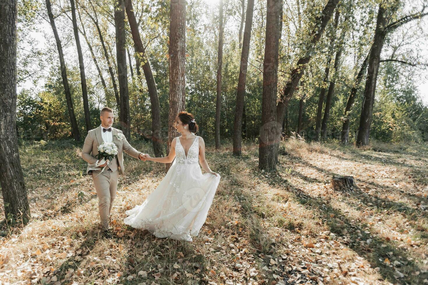 Wedding photo in nature. The bride and groom are walking in the forest, holding hands and looking at each other, the bride is holding her wedding dress beautifully. Couple in love. Summer light.