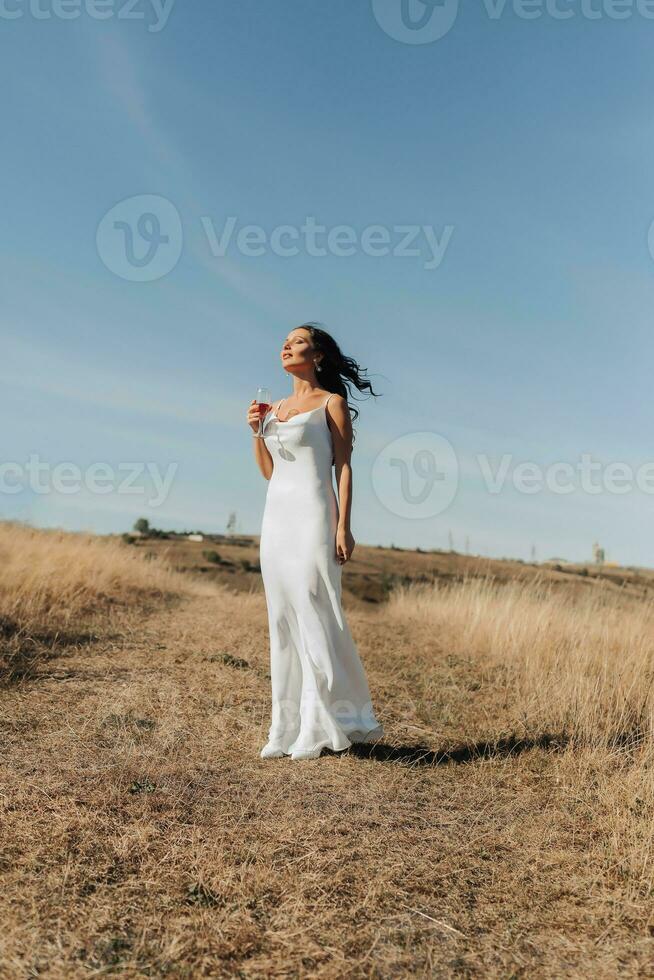 A girl with long, black, curly hair blowing in the wind is standing dressed in a white silk dress, holding a glass of champagne in her hand, against a background of blue sky and golden steppe. photo