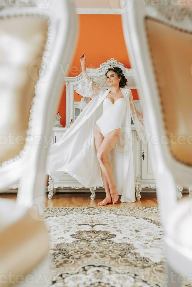 tall bride with bare shoulders, open bust, in a room near a mirror. Great wedding hairstyle. lace robe. Portrait. Delicate wedding makeup photo