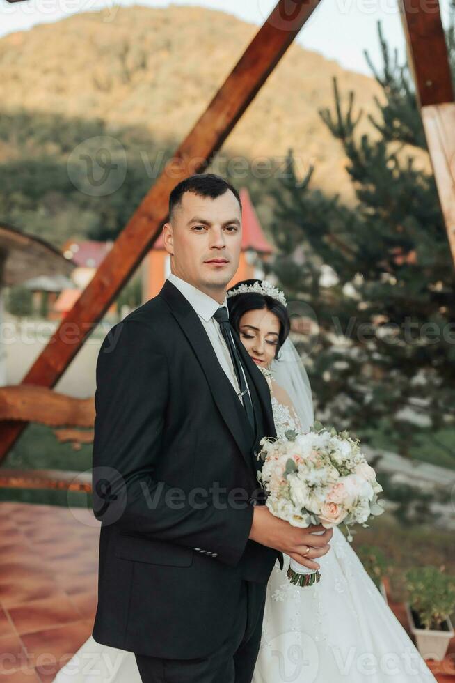 The brunette bride and groom in a white long dress are standing and hugging against the background of coniferous trees and a white hut. Stylish groom. Beautiful hair and makeup photo