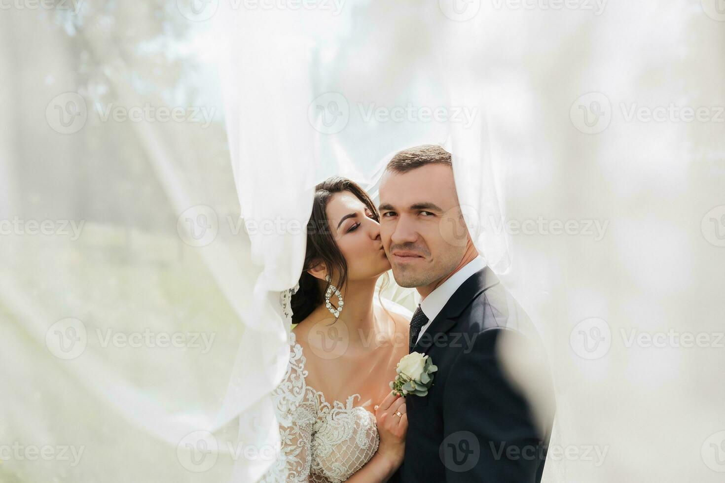 A curly-haired brunette bride in a white dress and a veiled groom embrace and kiss. Portrait of the bride and groom. Beautiful makeup and hair. Wedding in nature photo