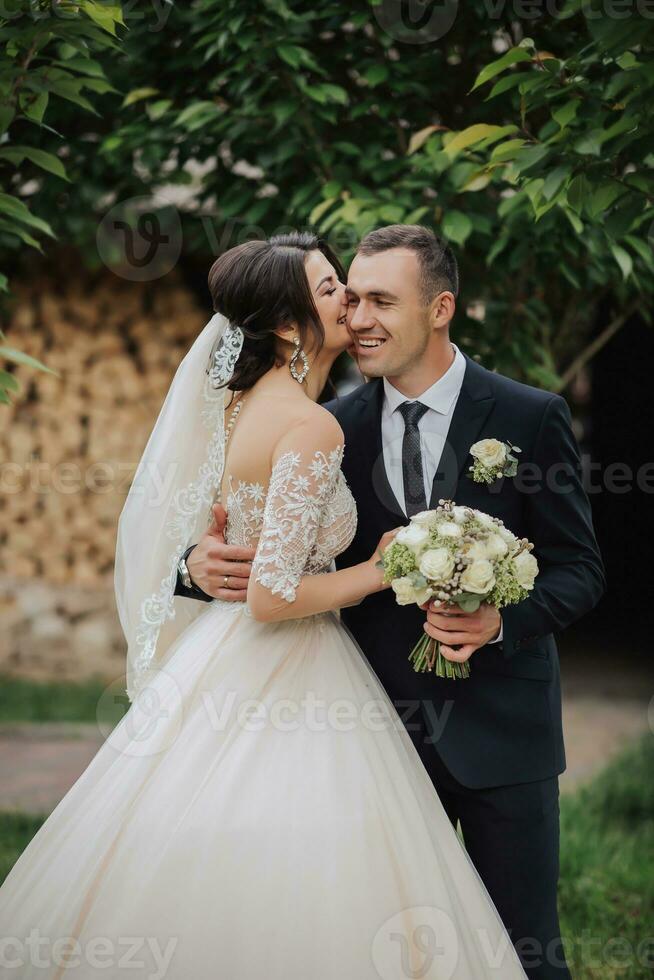 Portrait of the bride and groom in nature. Stylish bride and groom in a long lace dress are hugging and kissing near the trees in the garden. A happy couple in love photo