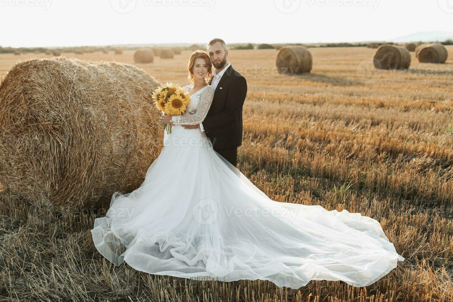 Wedding portrait of the bride and groom. The groom hugs the bride from behind, next to a bale of hay. Red-haired bride in a long dress with a bouquet of sunflowers. Stylish groom. Summer photo