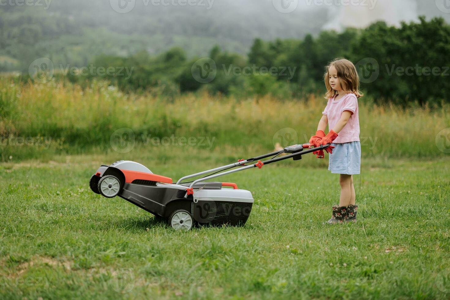 A child in boots in the form of a game mows grass with a lawnmower in the yard against the background of mountains and fog, the concept of garden tools photo