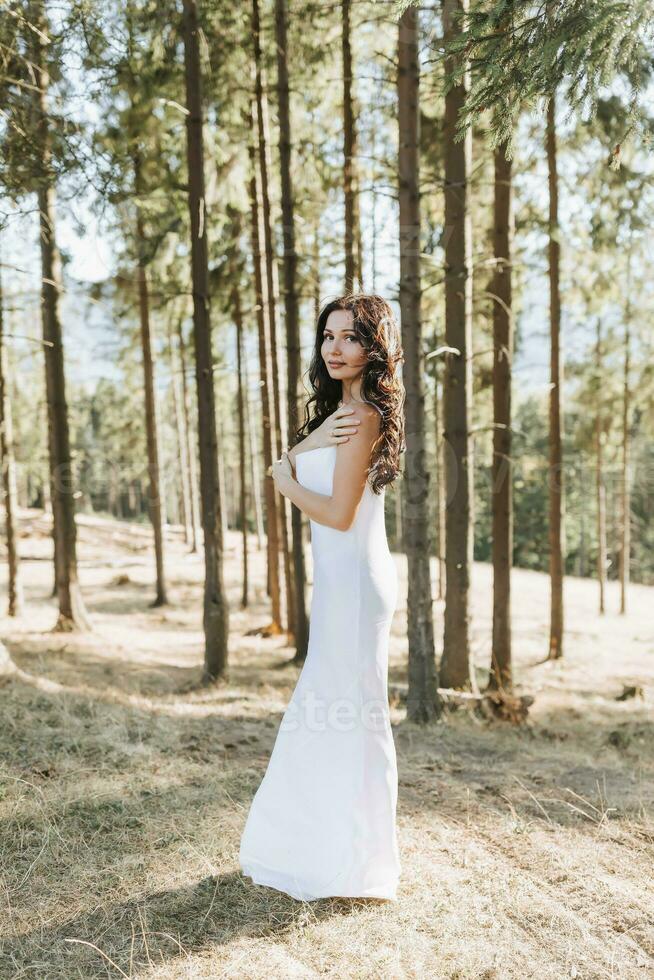 un niña con largo negro Rizado pelo soplo en el viento es en pie en un blanco seda vestido. en pie en el primavera bosque en contra el antecedentes de grande arboles foto