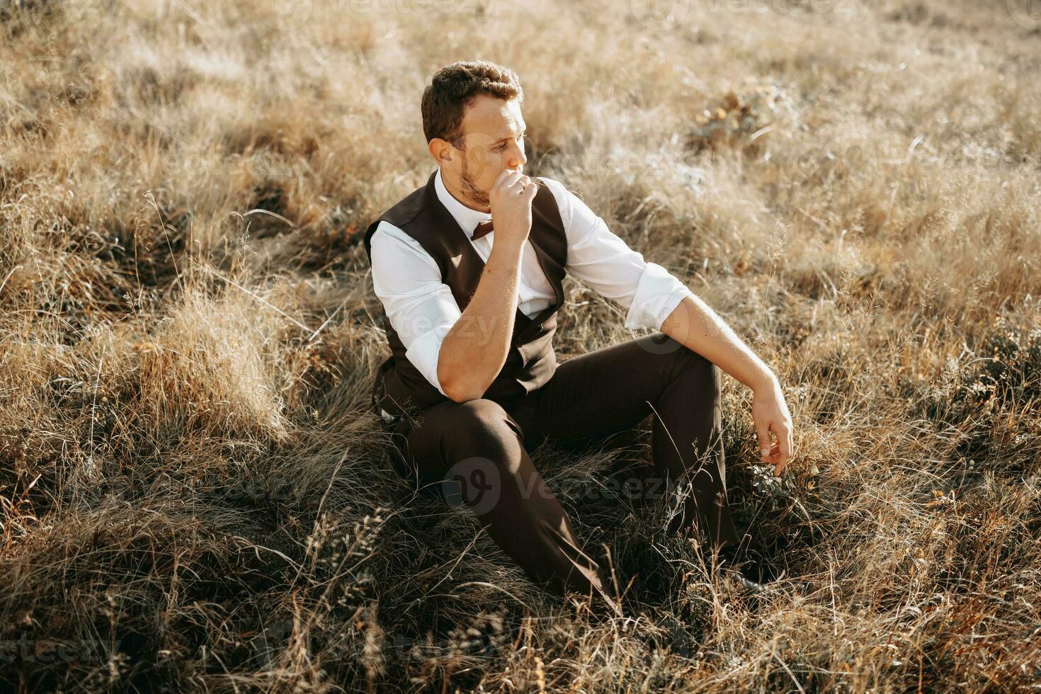 portrait of a stylish groom on a background of dry autumn grass. the concept of a rural wedding in the mountains, happy bohemian newlyweds. man relaxing sitting on the grass photo