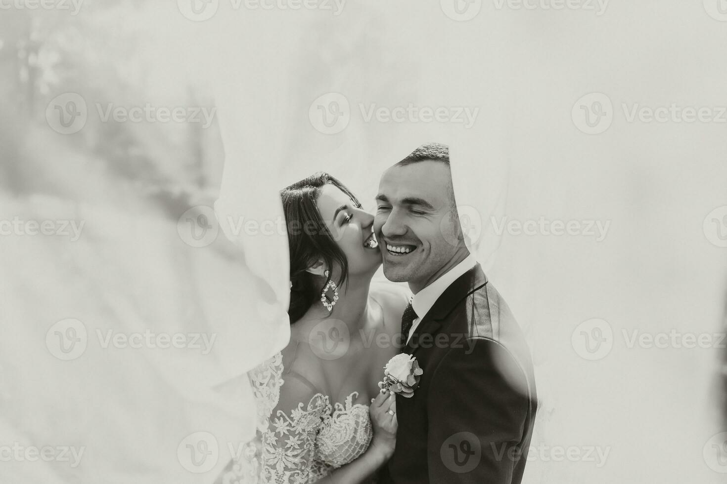 A curly-haired brunette bride in a white dress and a veiled groom embrace and kiss. Black and white Portrait of the bride and groom. Beautiful makeup and hair. Wedding in nature photo