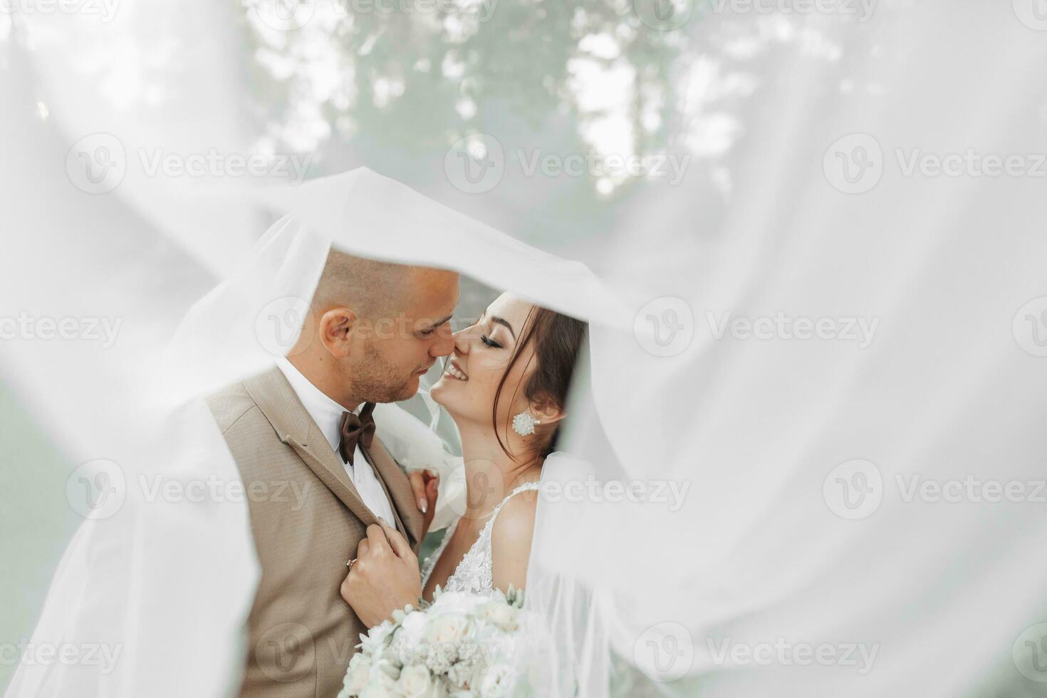 Wedding portrait of the bride and groom. Happy bride and groom gently hug each other under the veil, pose and kiss. Stylish groom. Beautiful young bride photo