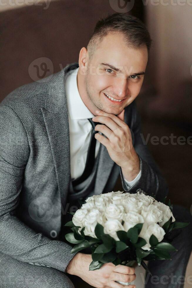 Portrait. A man in a white shirt, black tie and gray suit is sitting on a sofa, holding a bouquet of white roses. A stylish watch. Men's style. Fashion. Business photo
