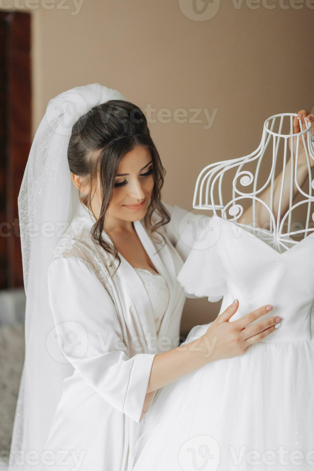 Brunette bride in a robe, posing for the camera, looking at her wedding  dress. Elegant hairstyle. Nice makeup. Voluminous veil. Morning of the  bride 36425634 Stock Photo at Vecteezy