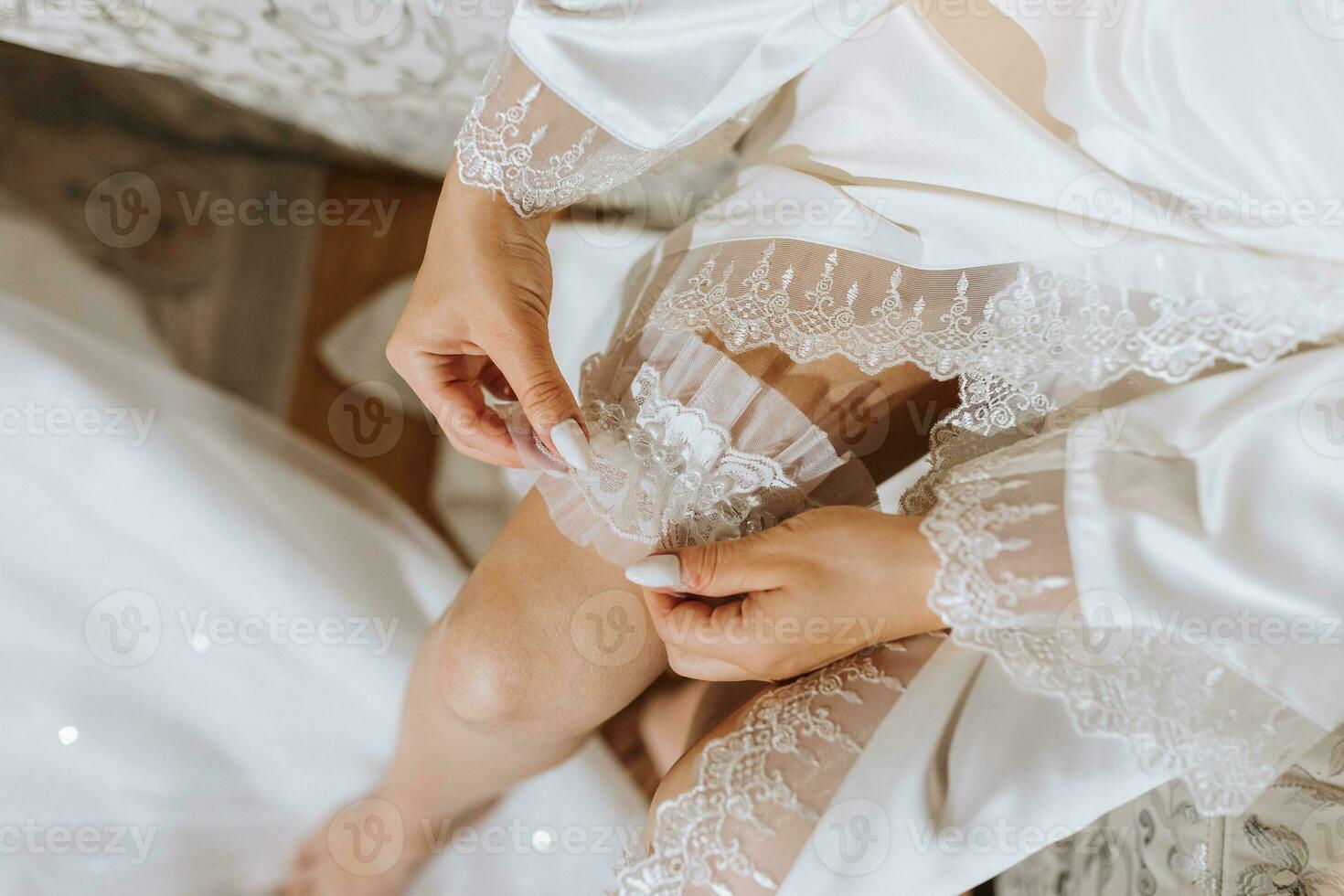 Bride dressed in a satin robe puts on a fishnet garter, close-up photo of hands. Beautiful hair and makeup. Wedding portrait. A beautiful bride in a dressing gown is posing