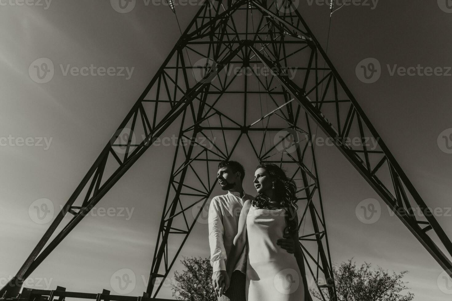 Stylish model couple in the mountains in summer. A young boy and girl in a white silk dress are standing near large structures of power lines. Black and white contrast photo